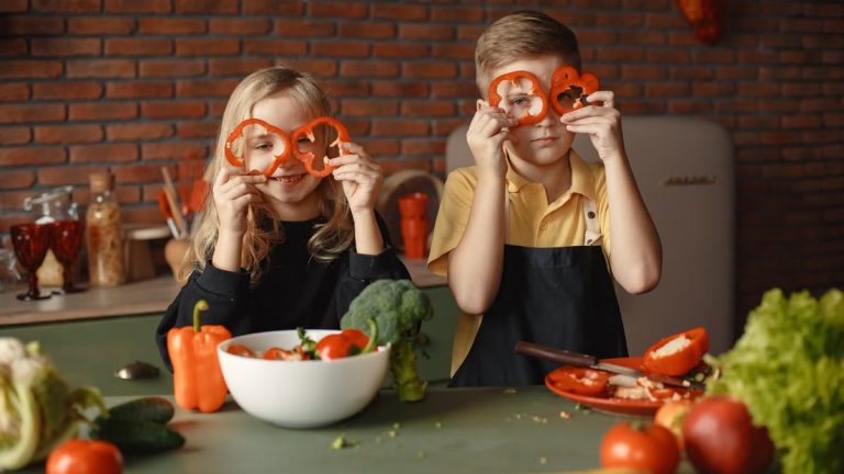 Funny boy and girl wearing apron looking through slices of pepper while preparing healthy vegetarian salad together at table with ingredients and bowls